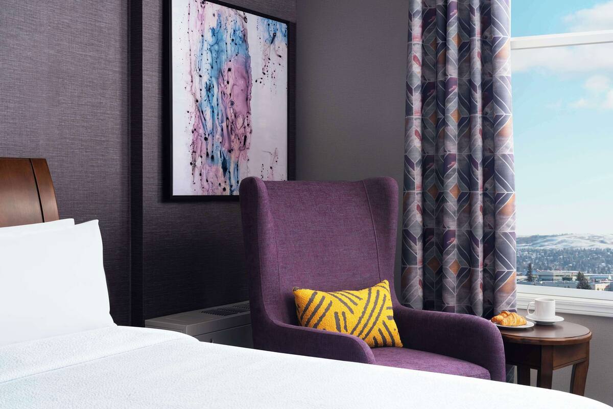 Habitación de hotel con una cama grande, un sillón púrpura con un cojín amarillo, un cuadro abstracto enmarcado y vistas a un paisaje urbano a través de una ventana con cortinas estampadas.