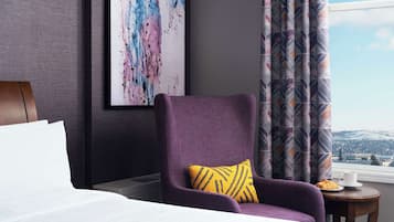 A hotel room with a large bed, a purple armchair with a yellow pillow, a framed abstract painting, and a view of a cityscape through a window with patterned curtains.