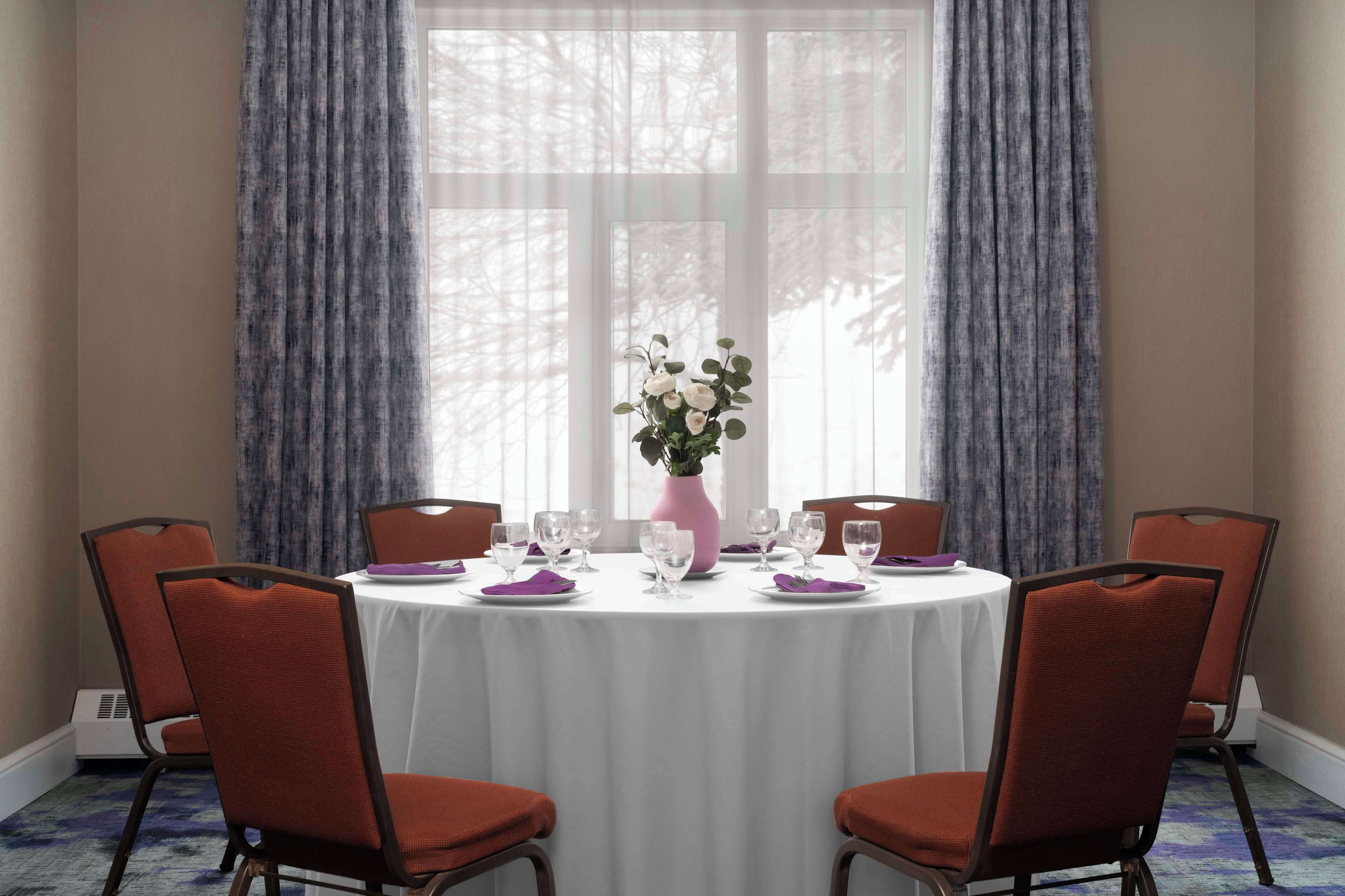A round dining table with a white tablecloth, surrounded by four red upholstered chairs. A vase with white flowers is placed in the center.