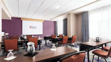 Ein Konferenzraum mit mehreren Tischen und Stühlen, einer Projektionsleinwand und dem Logo eines Hilton Garden Inn an der Wand.