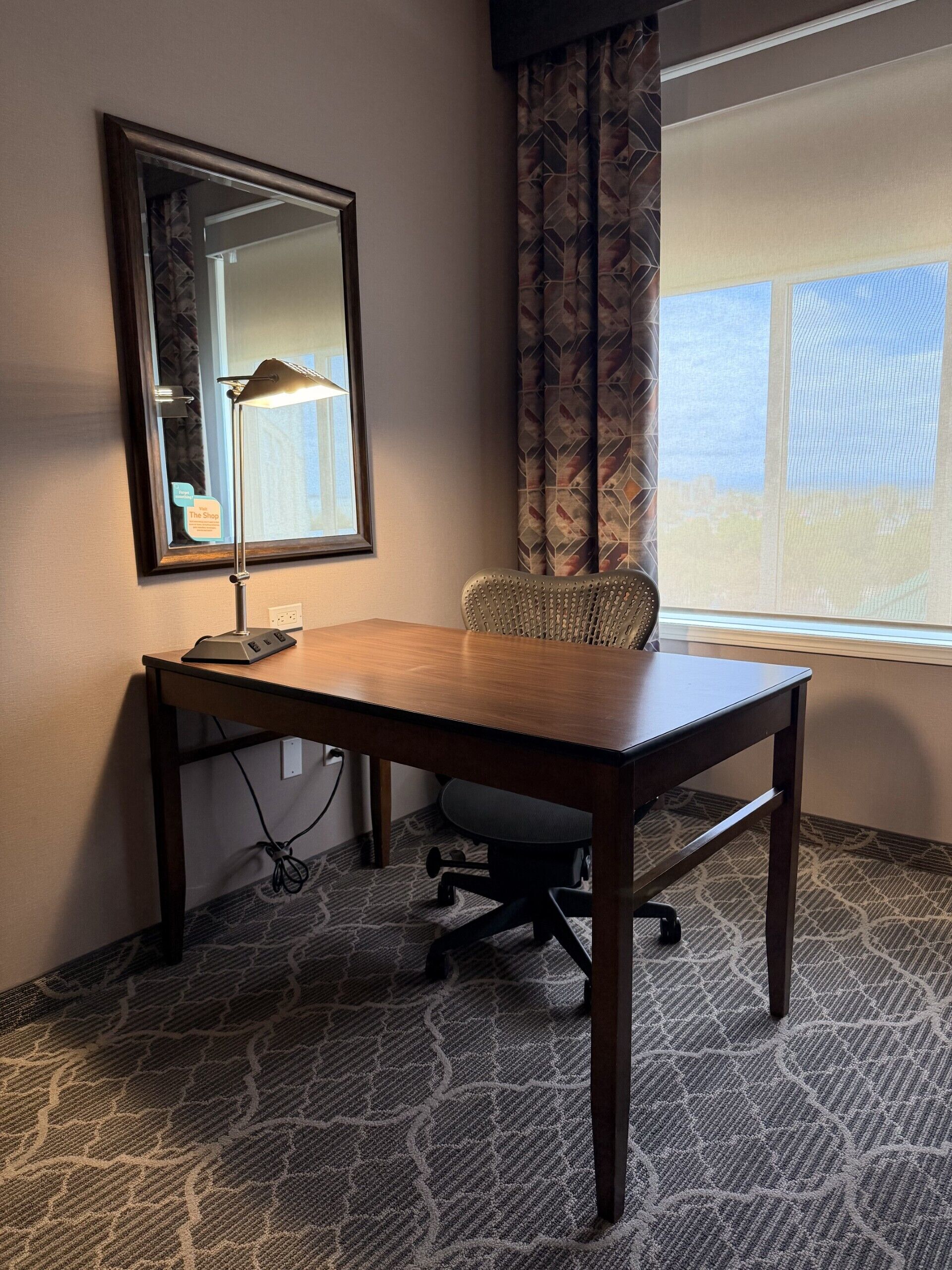 A hotel room with a wooden desk, a chair, a lamp, a mirror, and a window with curtains.