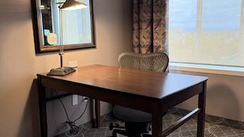 A hotel room with a wooden desk, a chair, a lamp, a mirror, and a window with curtains.