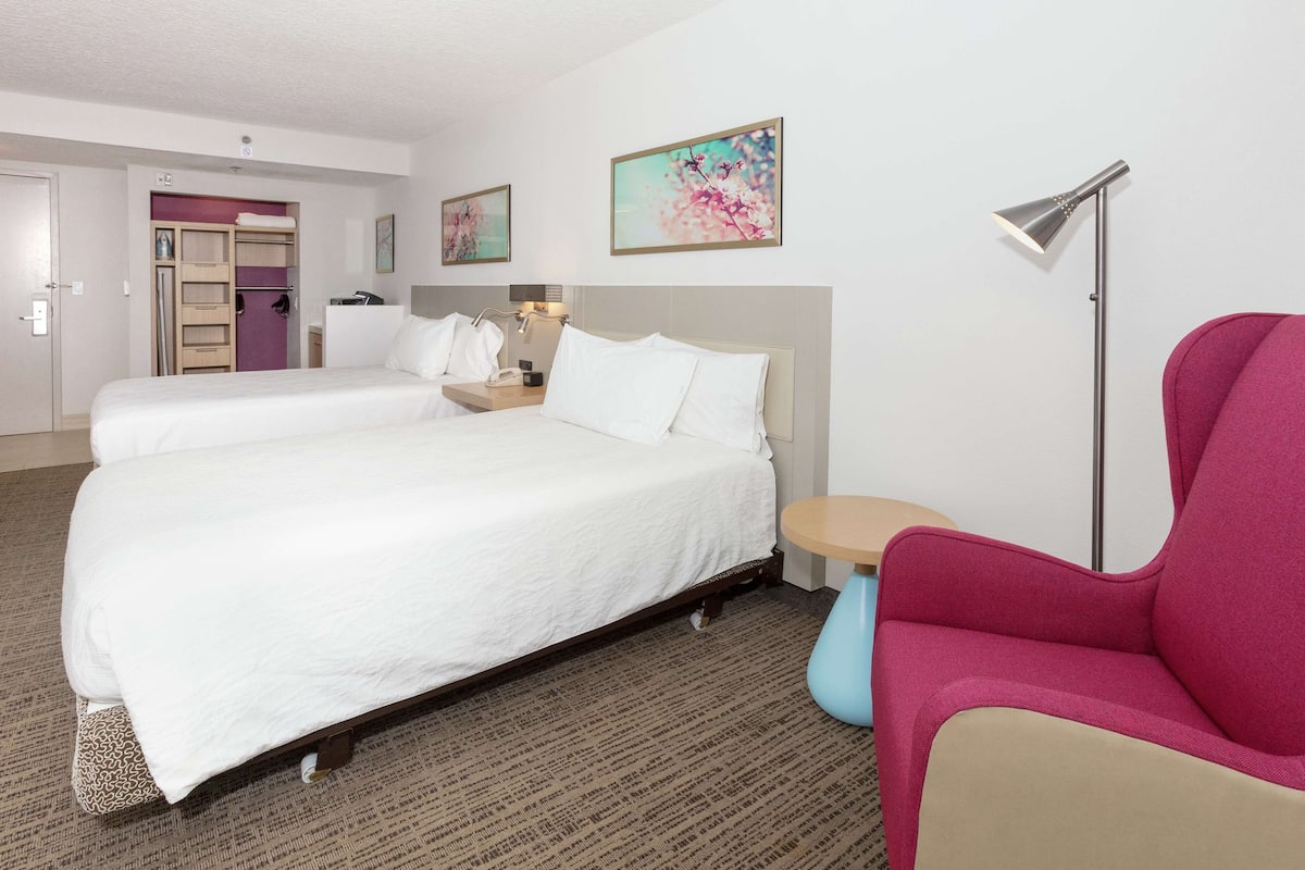 A hotel room with two beds, a desk, a chair, and a floor lamp.