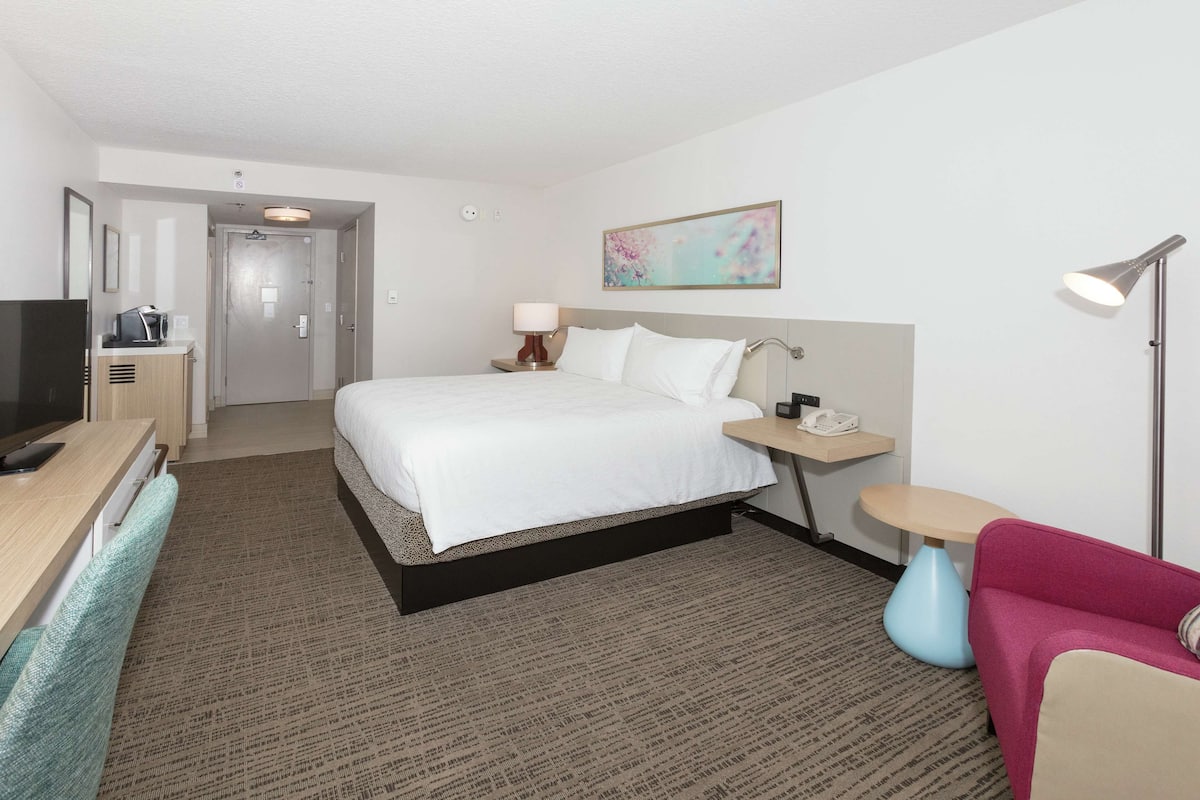 A hotel room with a bed, bedside table, lamp, TV, and a desk with a chair.