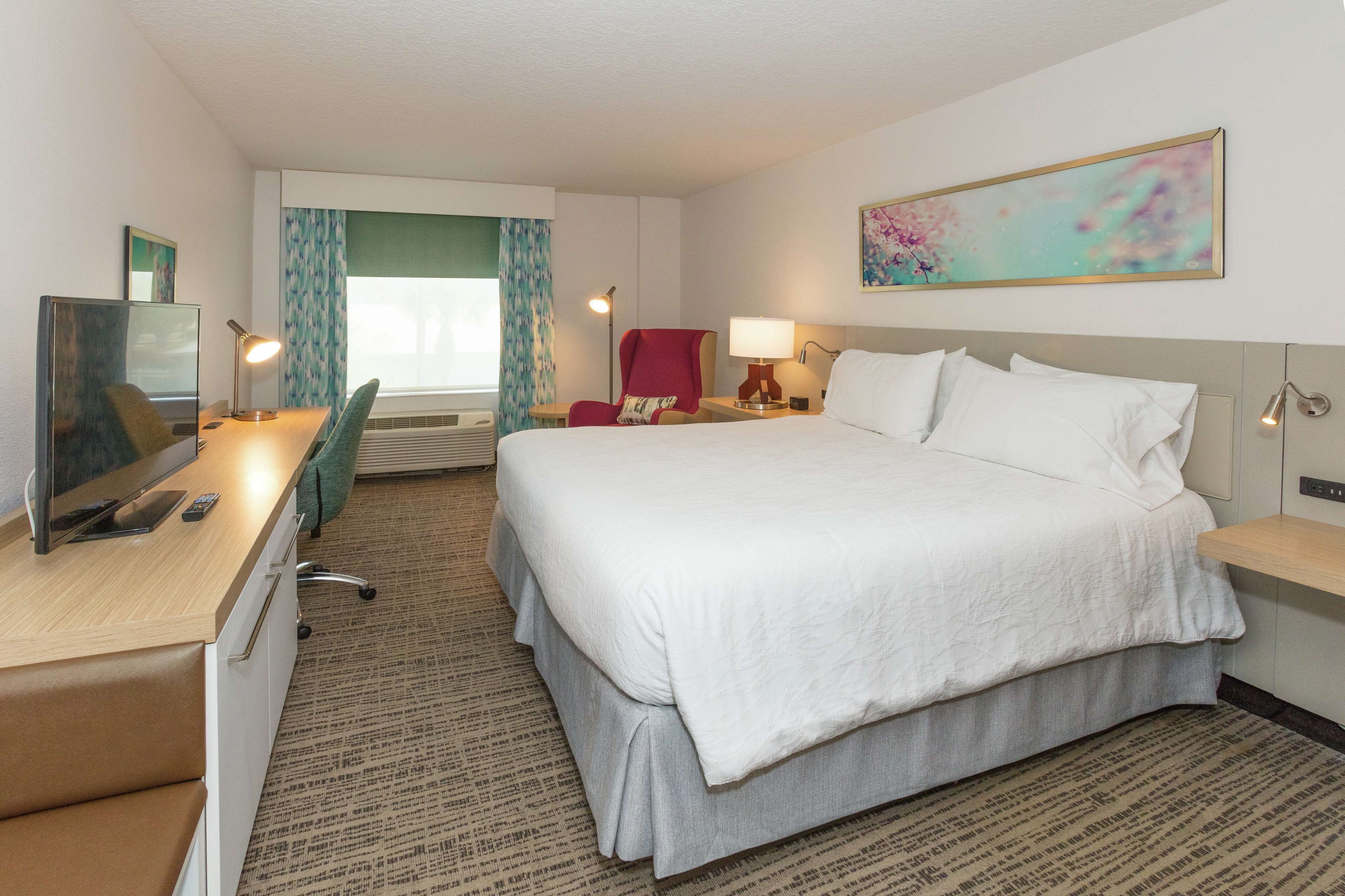 A hotel room with a bed, desk, chair, TV, and artwork on the wall.