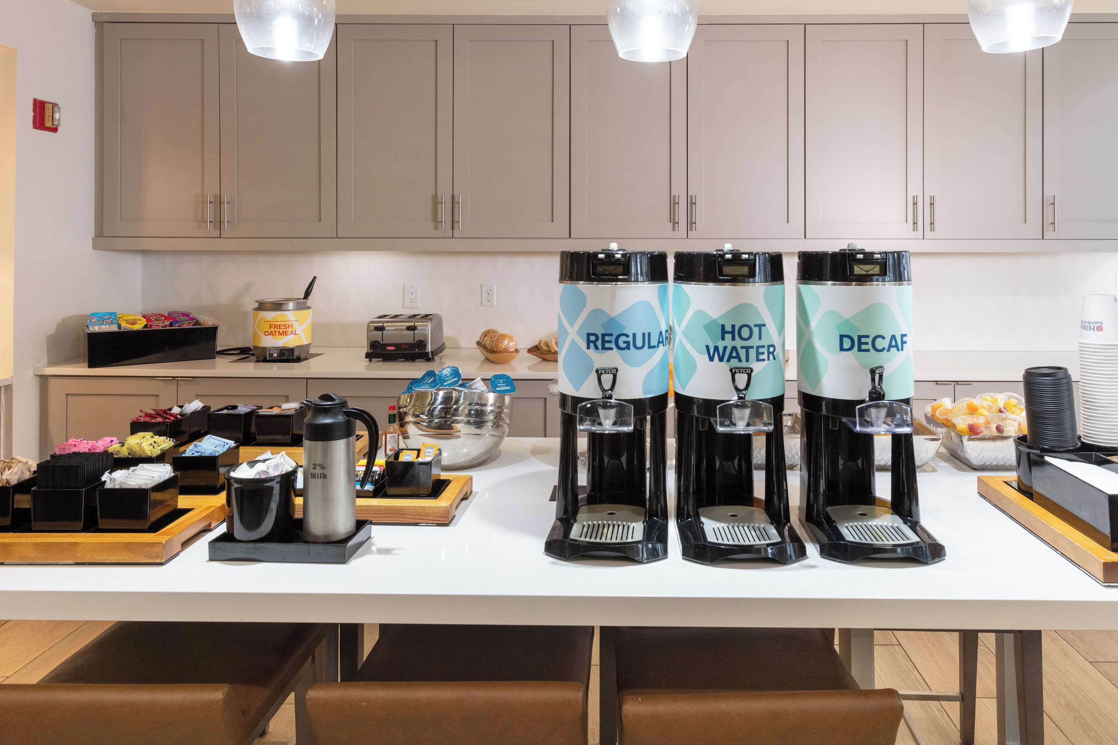 Un coin cuisine comprend un espace dédié au café, avec trois distributeurs de café étiquetés « Café normal », « Eau chaude » et « Café décaféiné ». On y trouve également divers aliments et un grille-pain sur le comptoir.