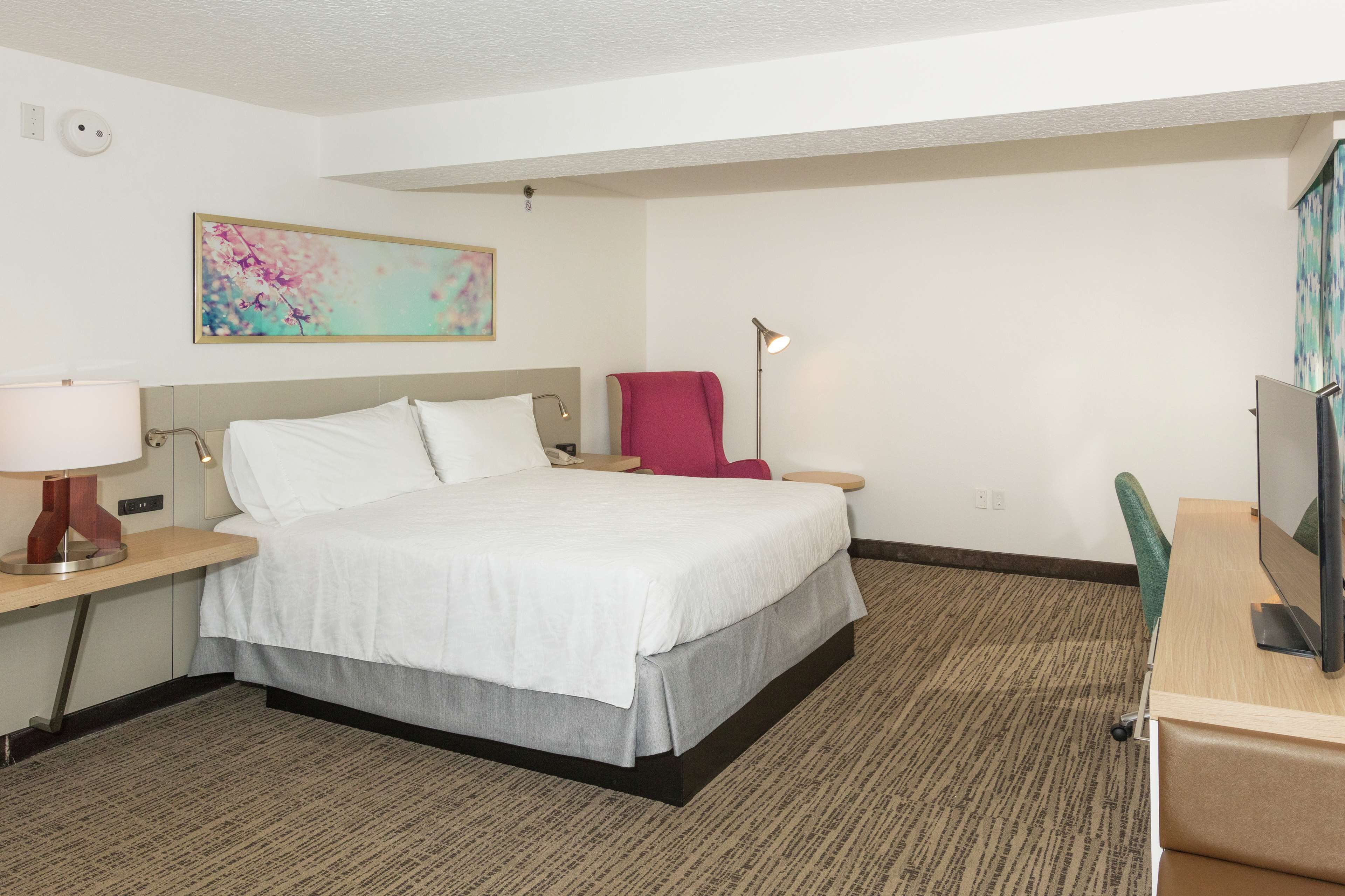 A hotel room with a bed, a desk with a computer, a chair, a lamp, and a painting on the wall.
