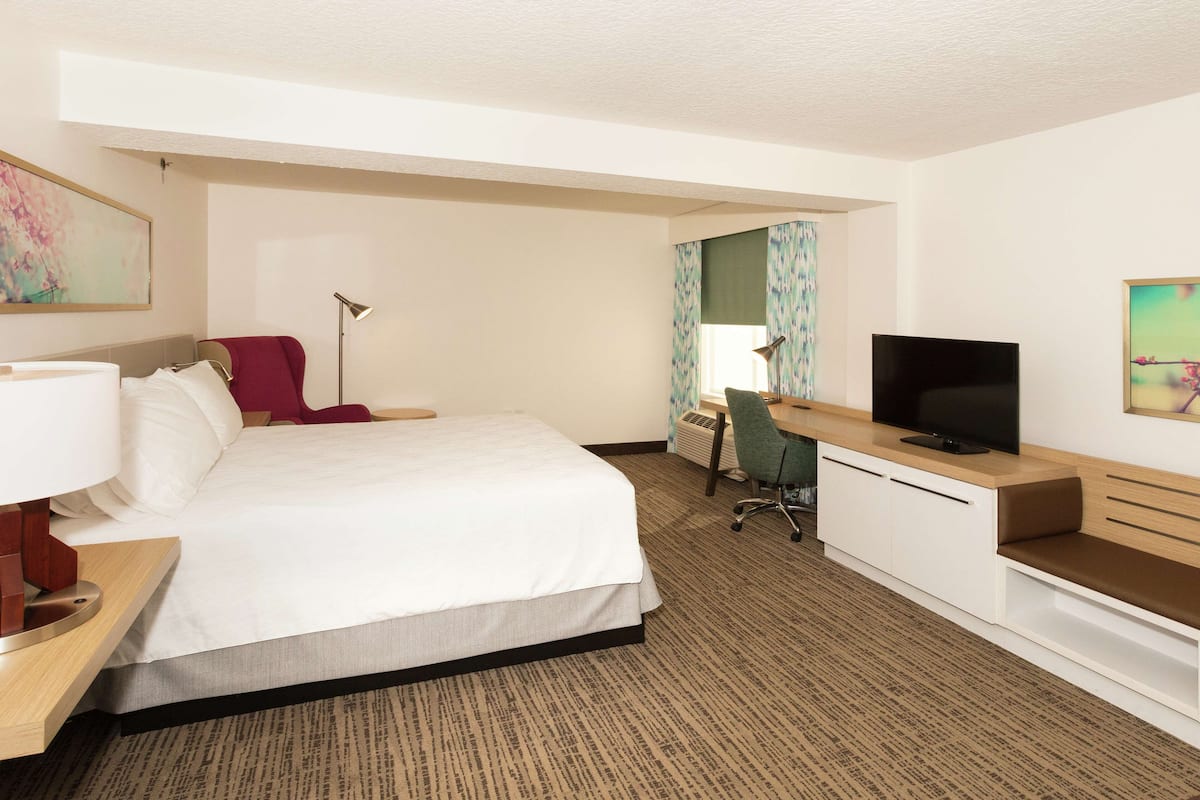 A hotel room with a bed, desk, TV, and a chair.