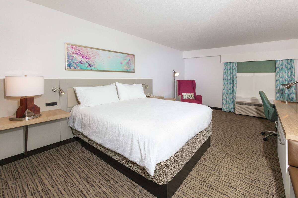 A hotel room with a large bed, a desk with a lamp, a chair, and a window with curtains.