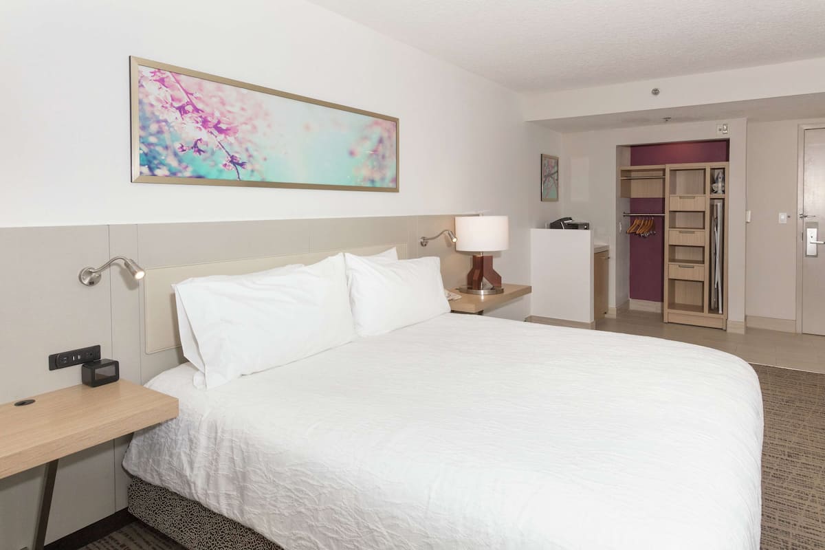 A hotel room with a large bed, a desk, a lamp, a wardrobe, and a painting on the wall.