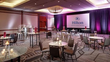 Ein geräumiger Konferenzraum mit runden Tischen und Stühlen, einem großen Bildschirm mit dem Hilton-Logo und einer Kristallleuchter.
