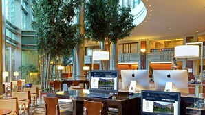 A modern hotel lobby with multiple computer stations, comfortable seating, and indoor plants.