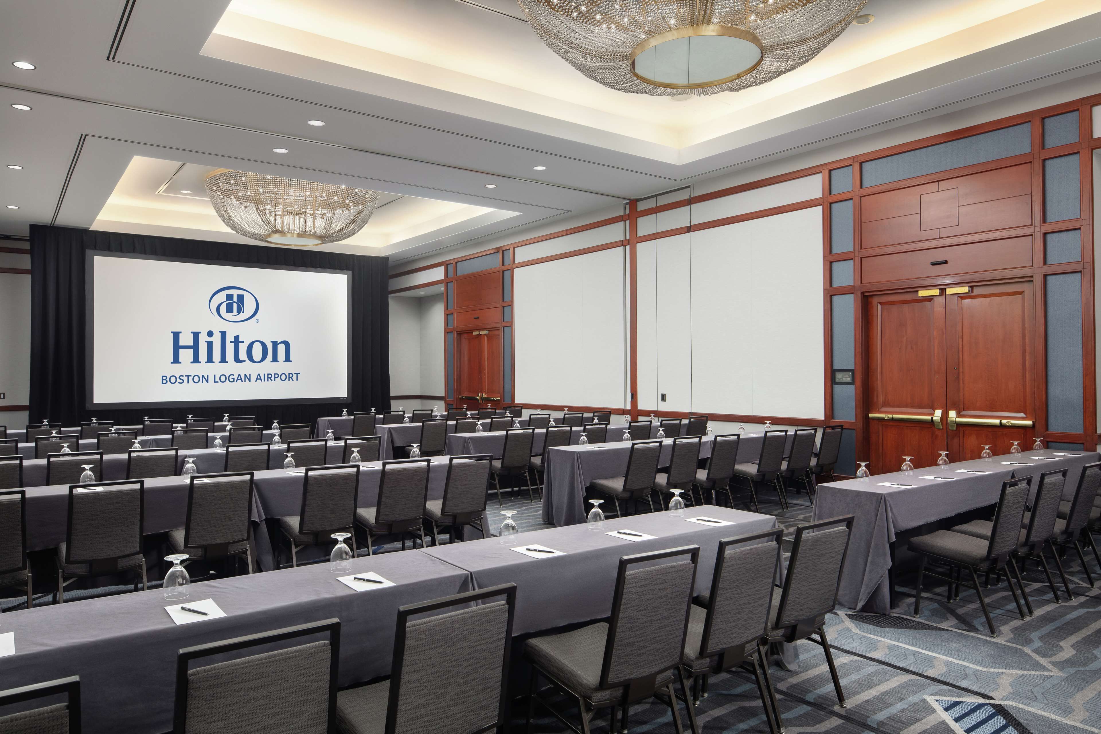 Una sala de conferencias con una pantalla grande que muestra 'Hilton Boston Logan Airport'. Filas de mesas con sillas están dispuestas para reuniones.