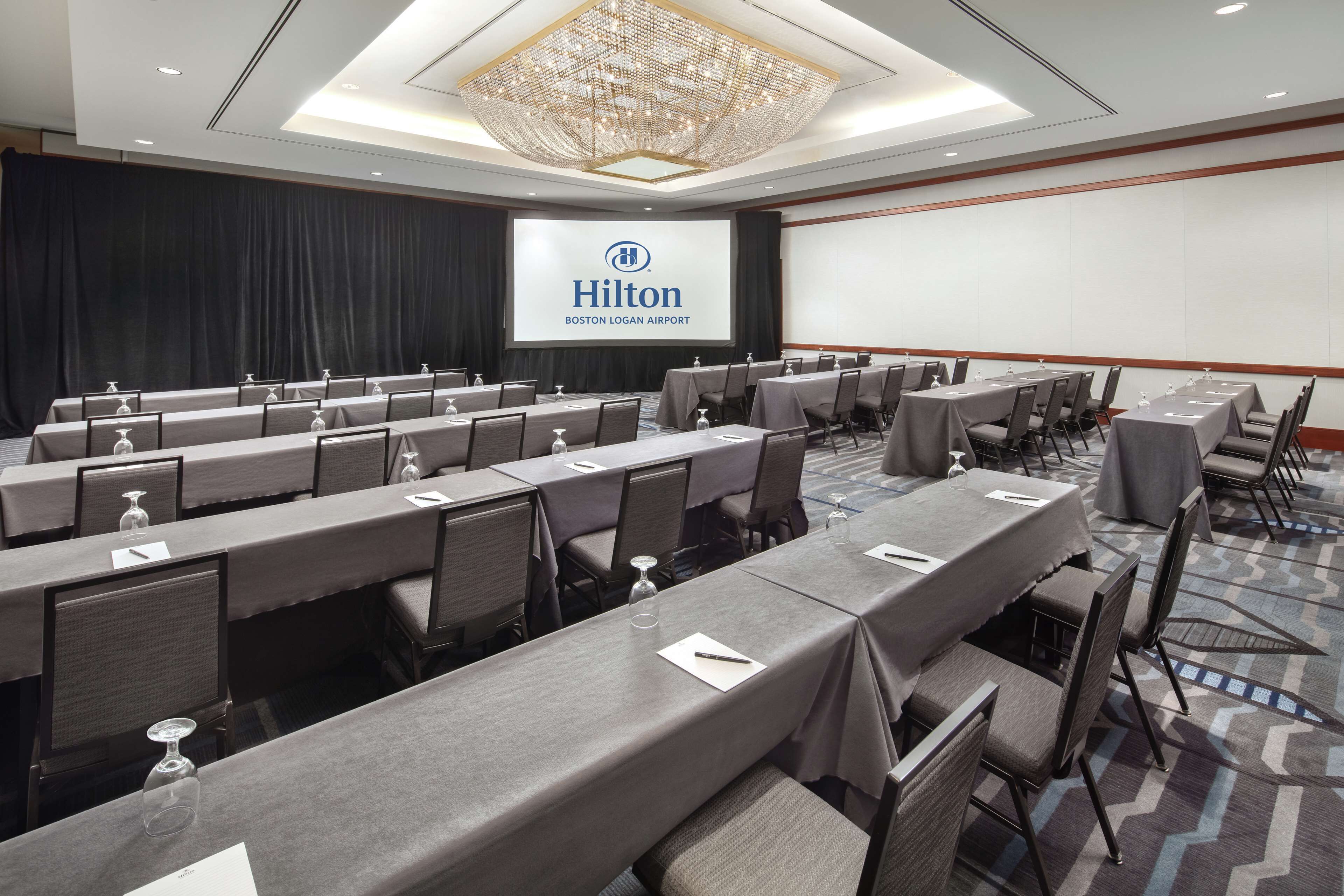 Un salón de conferencias con filas de mesas y sillas, una pantalla grande mostrando el logotipo de Hilton y una araña.
