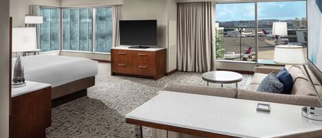 Habitación de hotel con cama, sofá, televisor, escritorio y vista al aeropuerto.