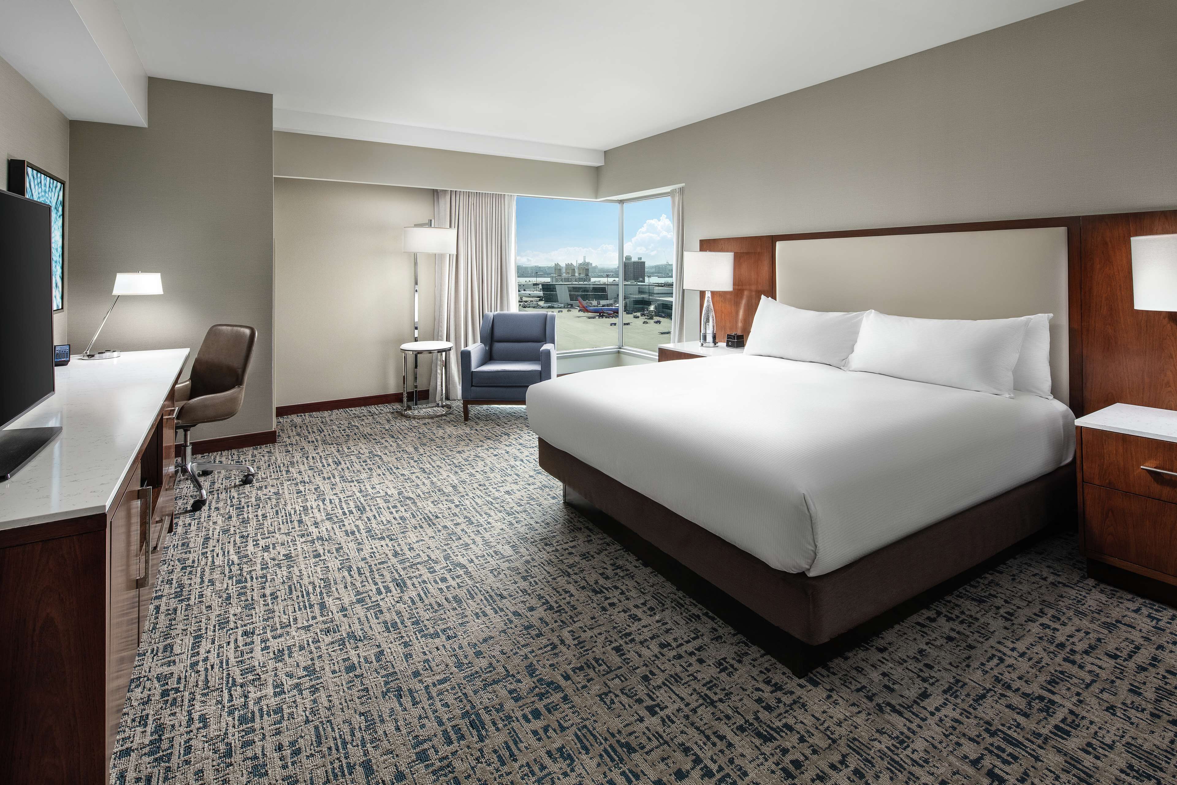 A hotel room with a large bed, a desk, a chair, and a view of the airport.