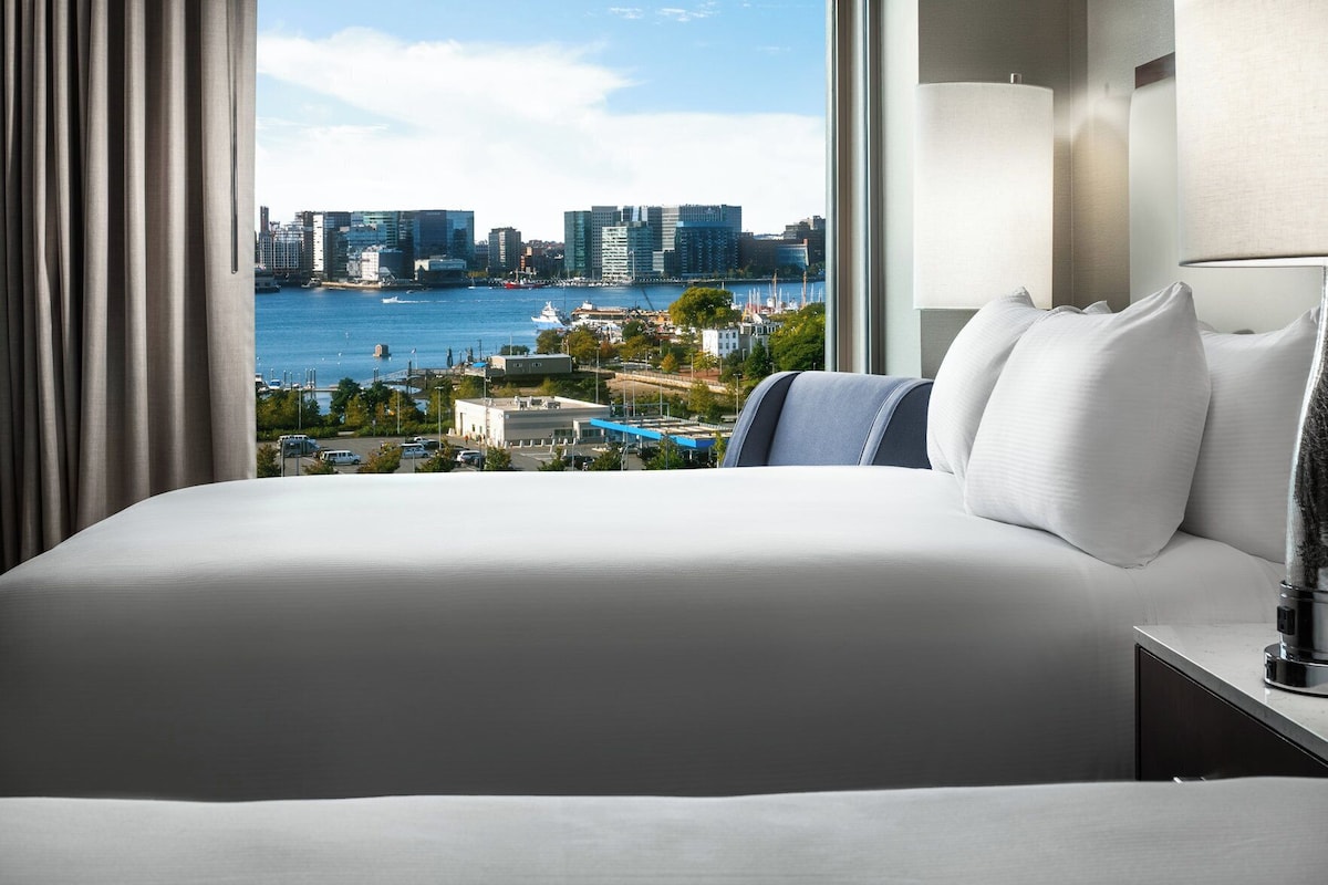 A hotel room with a large bed, a view of a cityscape and water, and a lamp on the nightstand.