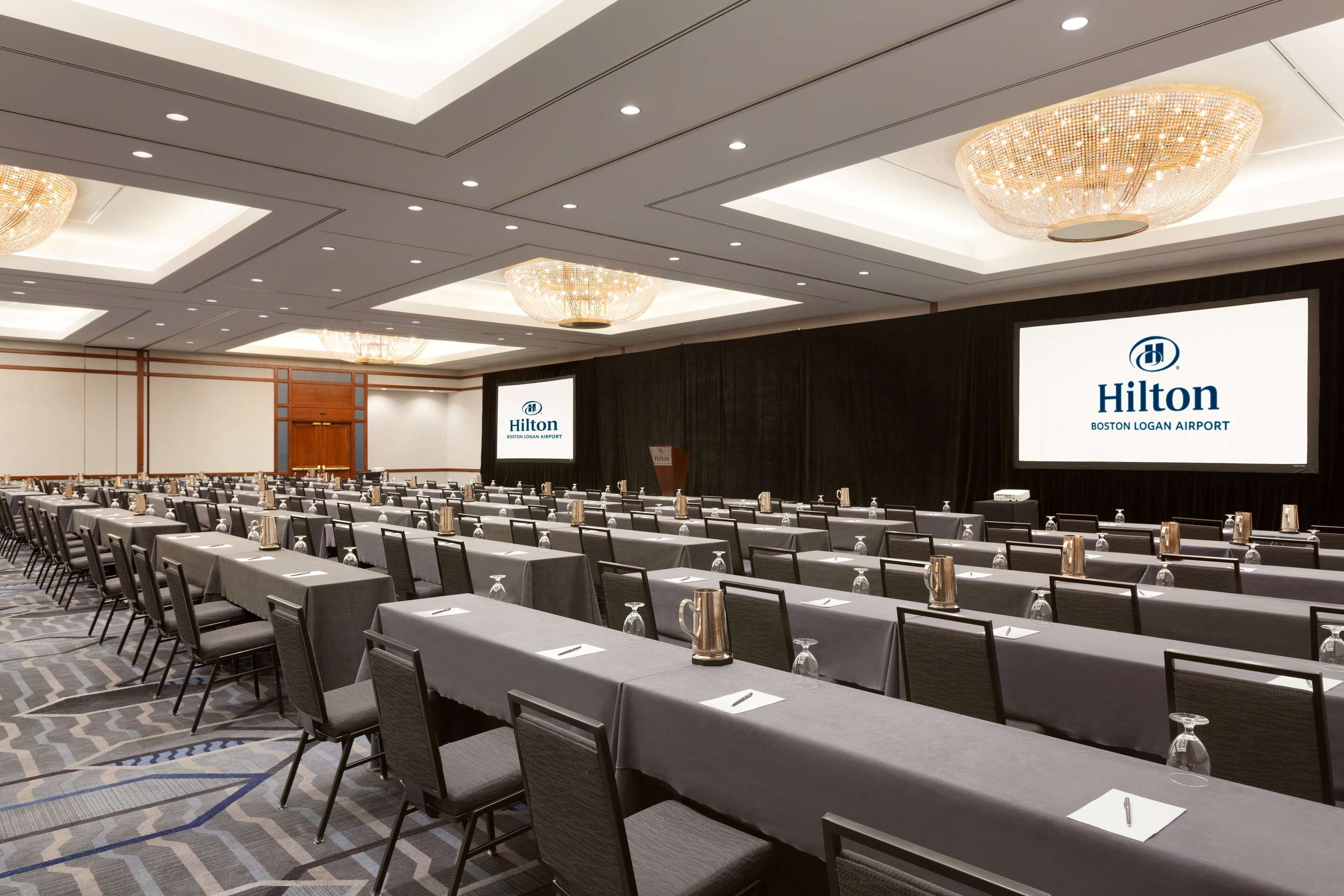 Ein Konferenzraum mit Tischen, Stühlen und einem großen Bildschirm, auf dem das Hilton-Logo zu sehen ist.