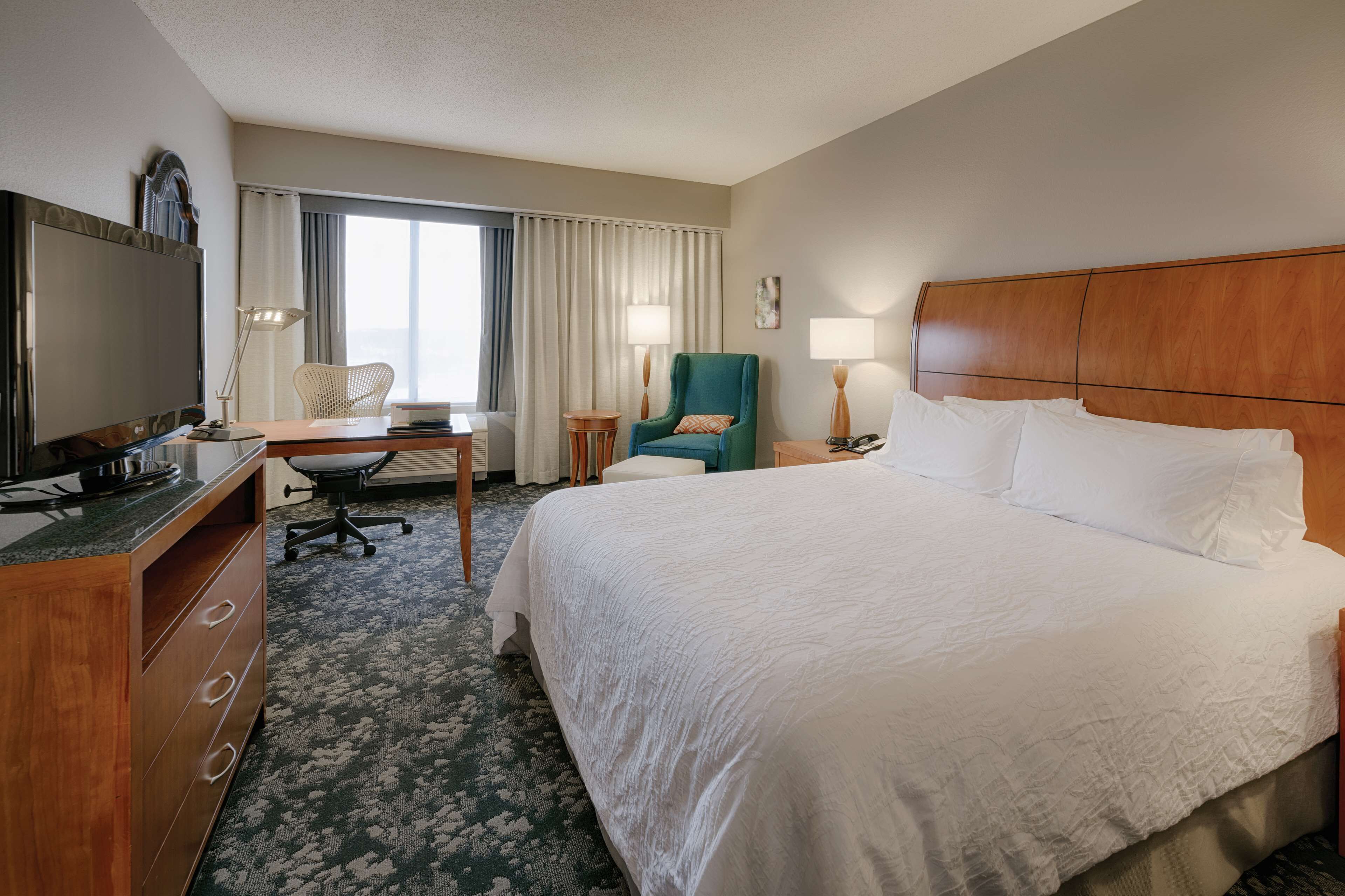 Une chambre d’hôtel avec un grand lit, un bureau avec un ordinateur, une chaise, une télévision et une fenêtre avec des rideaux.