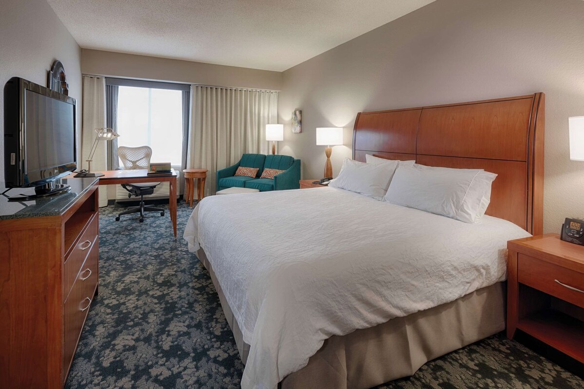 A hotel room with a large bed, a desk with a TV, a chair, and a lamp.