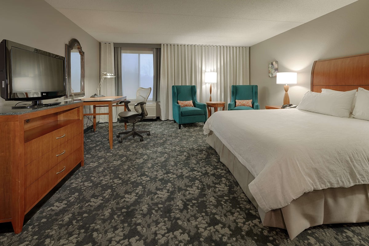 A hotel room with a bed, a desk with a computer, two chairs, a TV, and a lamp.
