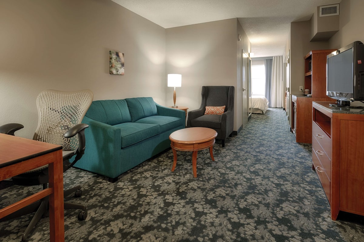 A hotel room with a teal sofa, a wooden side table, a grey armchair, a small round table, a TV, and a kitchenette area.