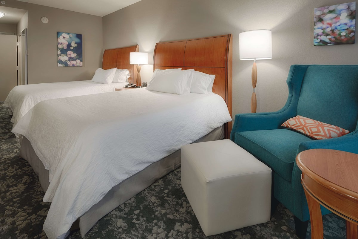 A hotel room with two beds, a teal armchair, a small round table, and a patterned carpet.