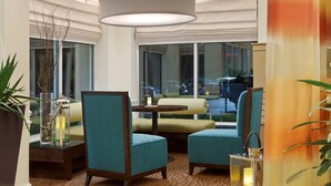 Um saguão de hotel com área de estar, com cadeiras verde-água, uma mesa redonda e uma janela ampla com vista para a rua.