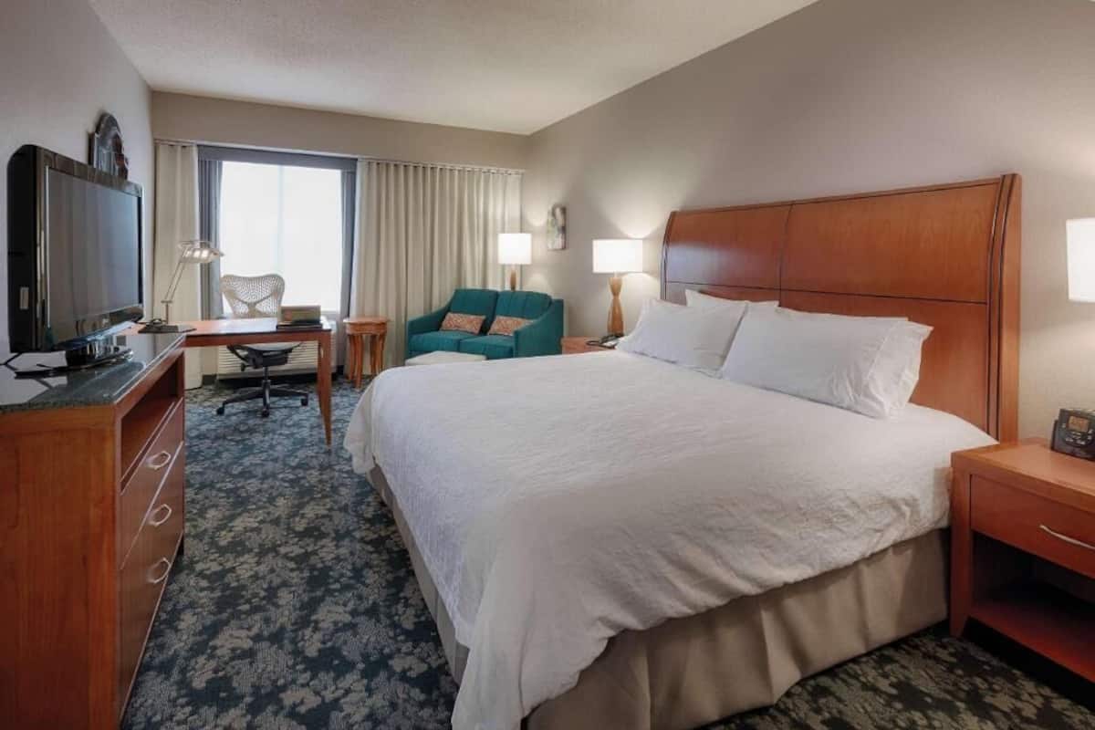 A hotel room with a large bed, a desk with a computer, a television, a chair, and a window with curtains.