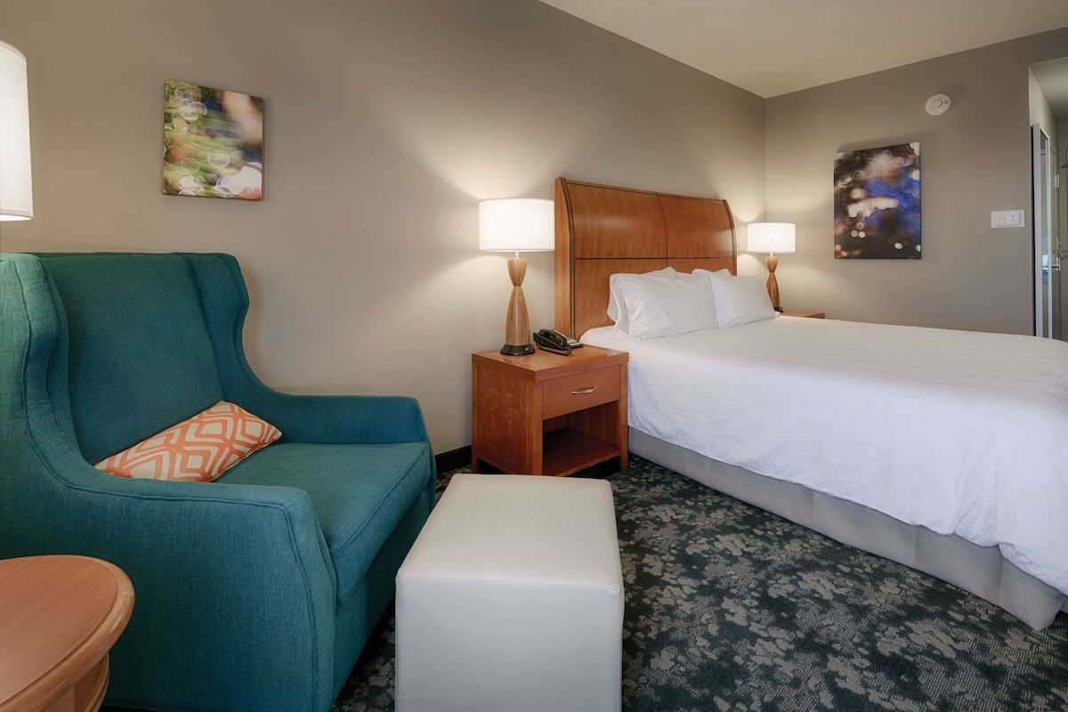 A hotel room with a bed, a green armchair, a bedside table with a lamp, and a small nightstand.