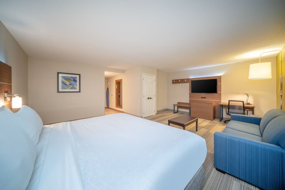 A hotel room with a large bed, a sofa, a desk, a TV, and a door leading to a bathroom.