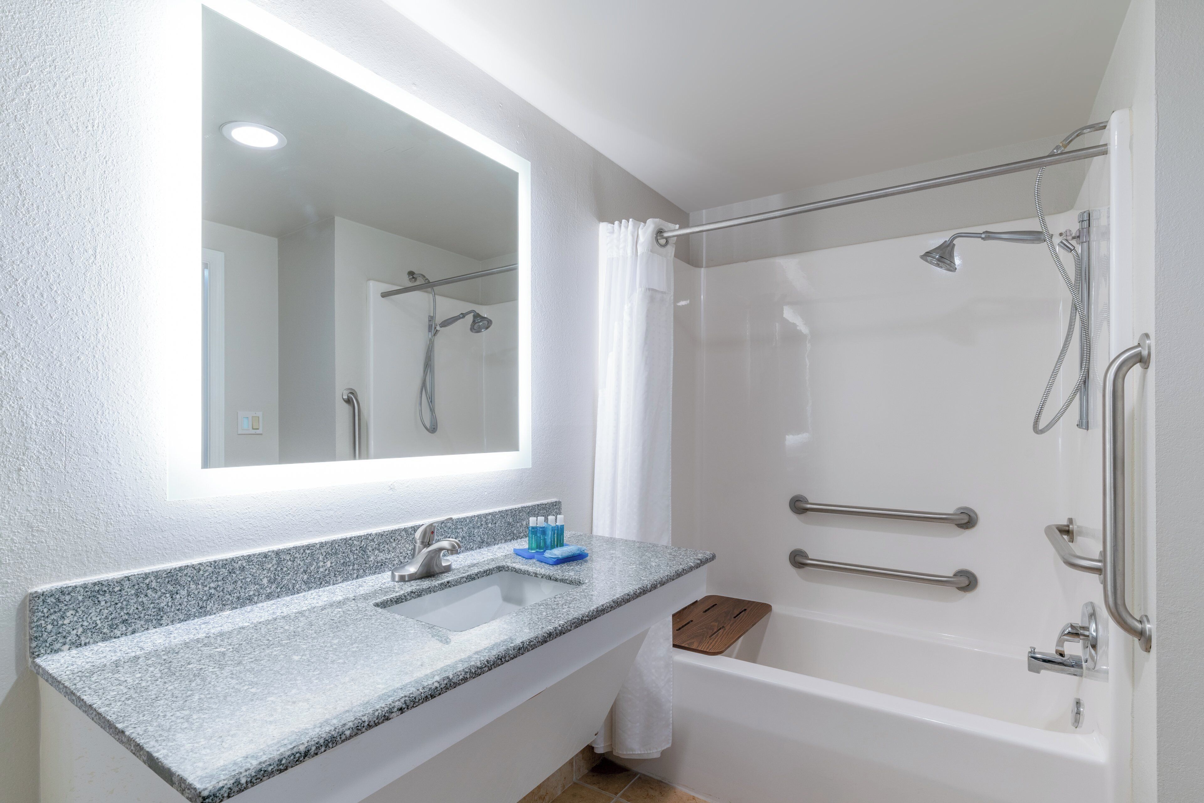 Un baño con encimera de granito, un espejo, ducha con barras de apoyo y una bañera blanca.