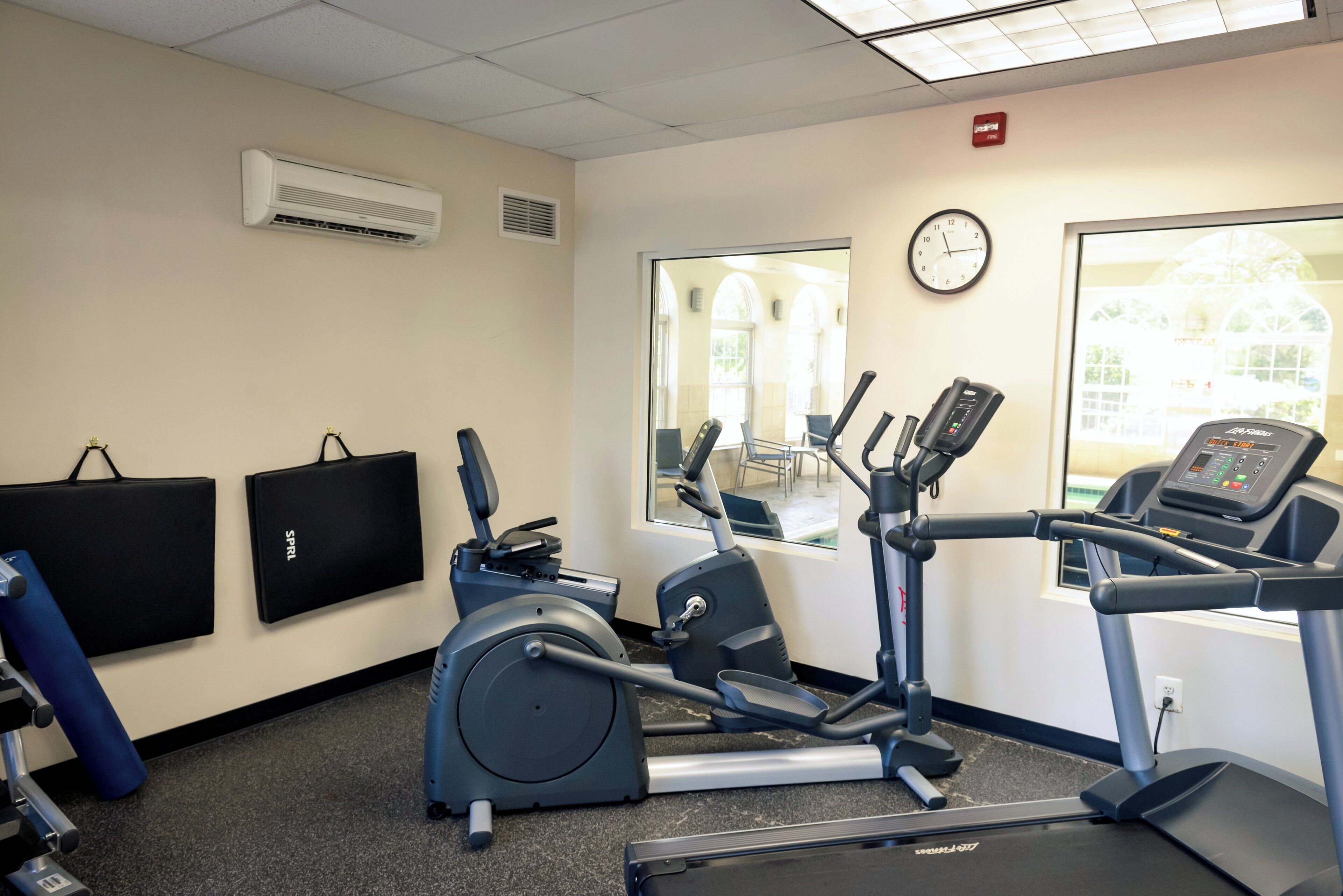 A fitness room with exercise equipment, a wall clock, and a window with a view of a building.