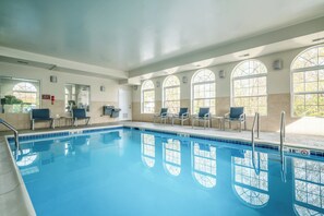 A swimming pool with clear blue water, surrounded by sunlit arched windows and a row of chairs.