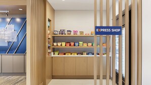 Um hall de hotel com um balcão de atendimento em madeira, uma placa azul com a inscrição "EXPRESS SHOP" e prateleiras cheias de lanches.