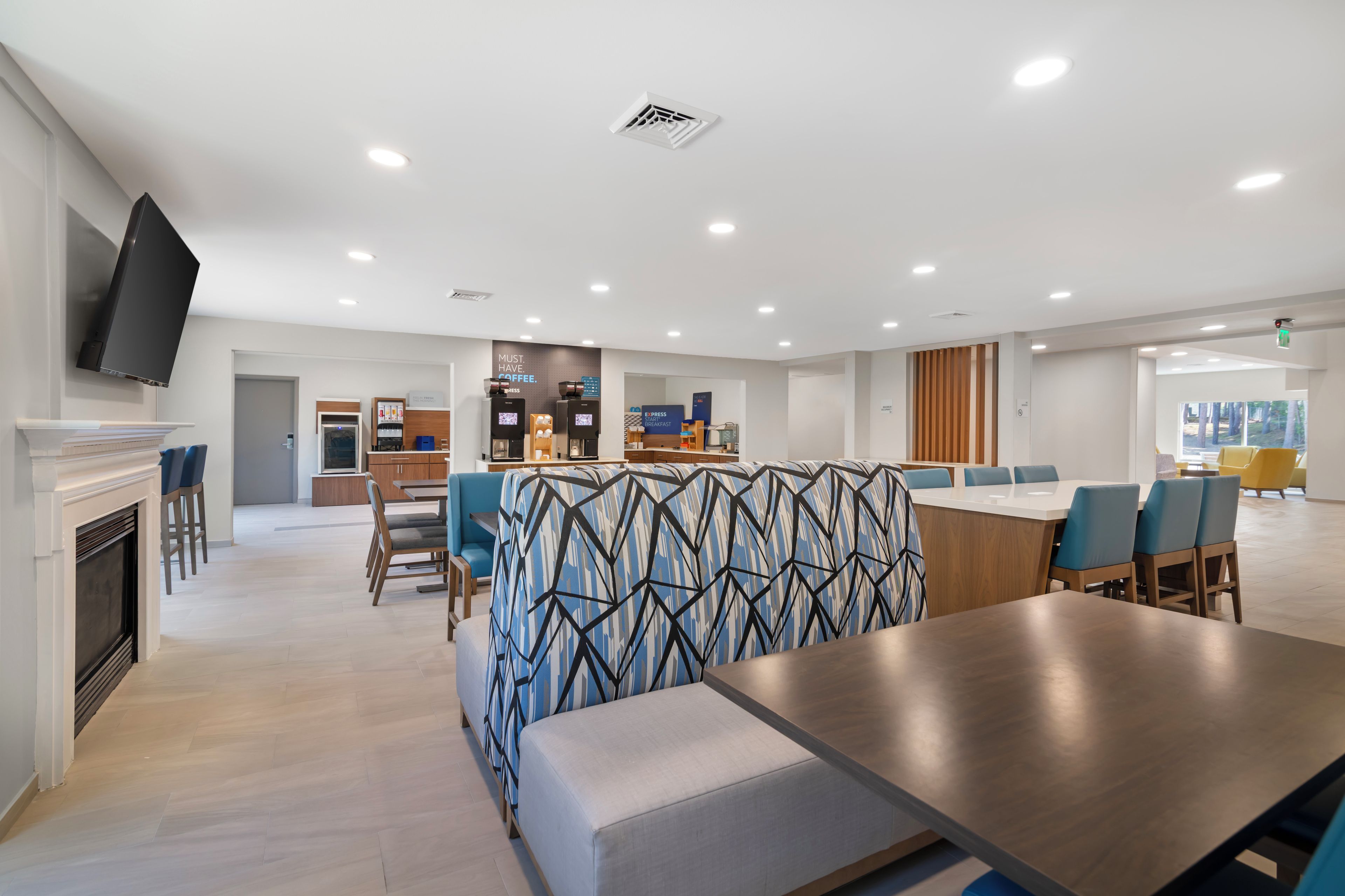 A modern dining area with a long table, blue upholstered seating, and a kitchen area in the background.