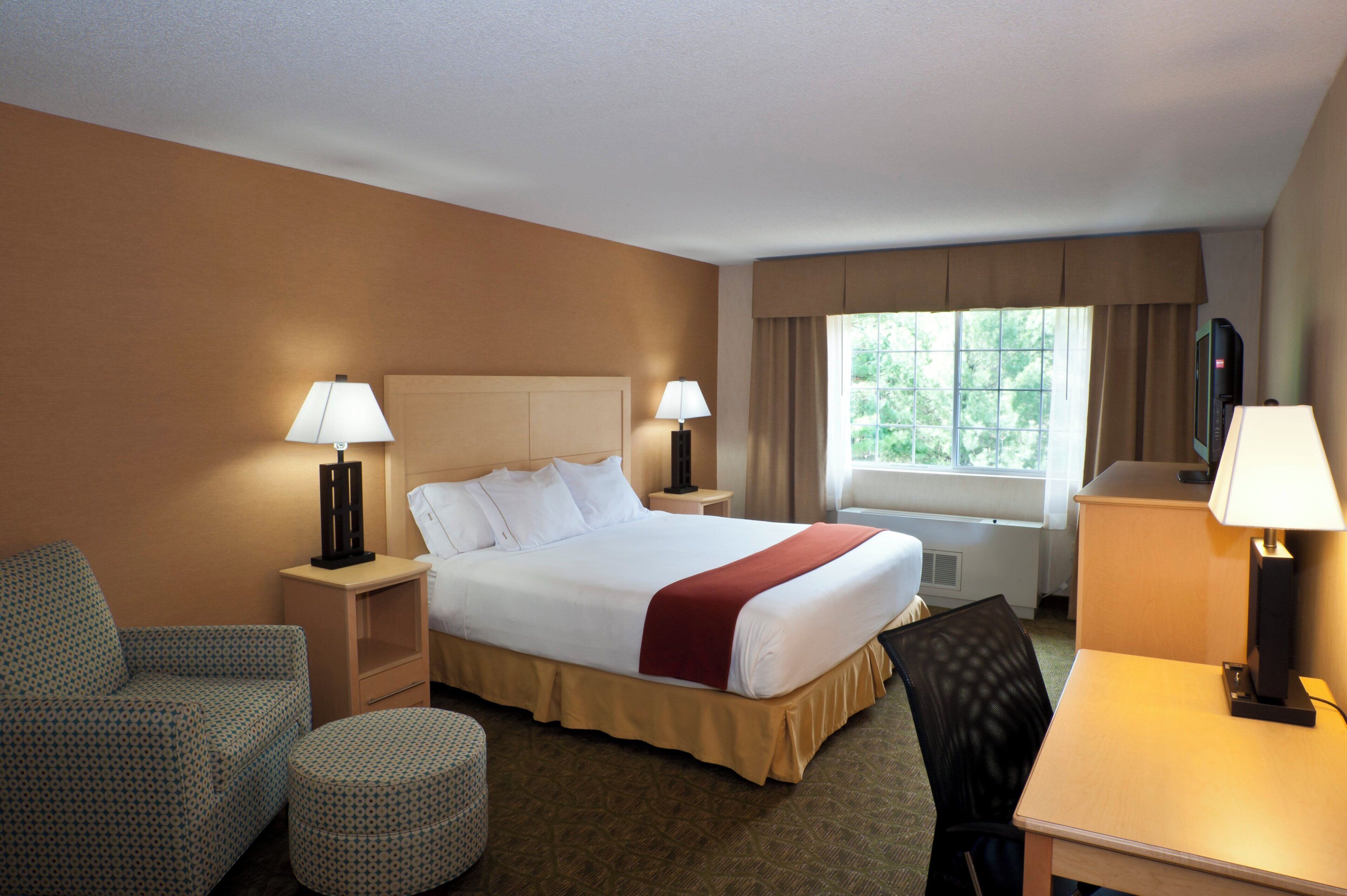 A hotel room with a bed, two bedside tables with lamps, a chair, a desk, and a window with curtains.