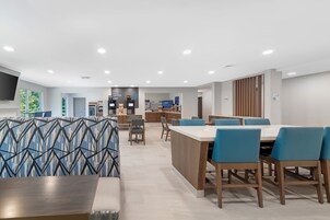 Un comedor moderno con una mesa larga y sillas azules, una cocina al fondo y un ventanal con vistas a zonas verdes.