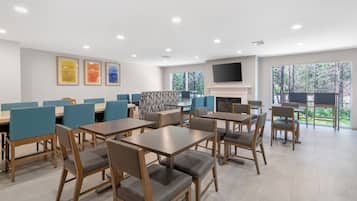 A modern dining area with multiple tables and chairs, a fireplace, and large windows offering a view of trees.