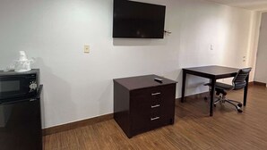 A room with a flat-screen TV mounted on the wall, a small wooden cabinet with drawers, a desk with a chair, and a microwave on the floor.