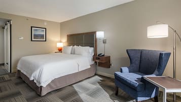 Habitación de hotel con una cama grande, una silla, un escritorio, una mesita de noche con lámpara y un cuadro en la pared.