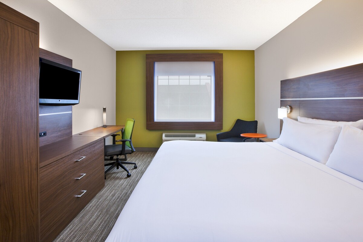 A hotel room with a bed, a desk with a flat-screen TV, a chair, a window, and an air conditioning unit.