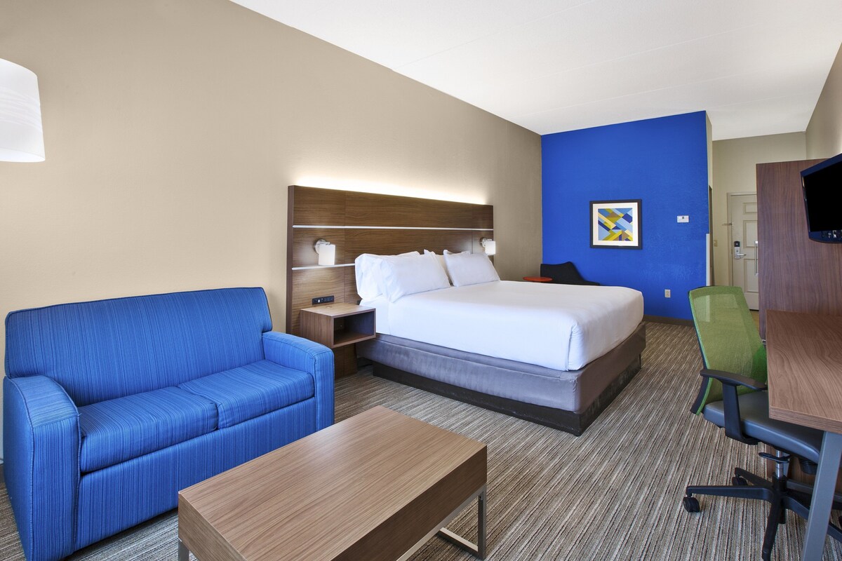 A hotel room with a bed, a blue sofa, a wooden coffee table, a desk with a chair, and a television.