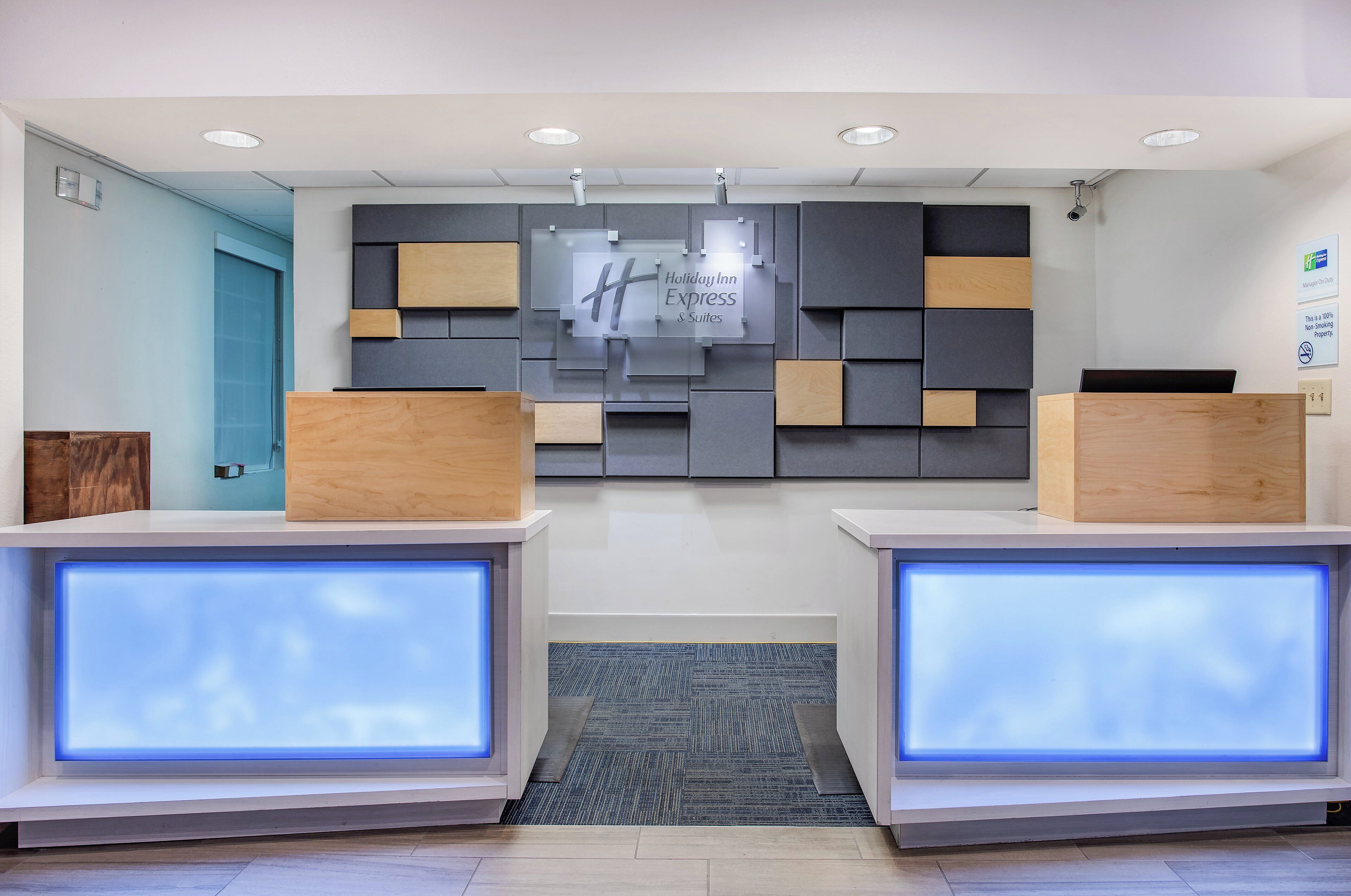 Un comptoir d’accueil d’hôtel au design moderne, avec une enseigne Holiday Inn Express, un panneau indiquant qu’il est interdit de fumer et une caméra de surveillance.