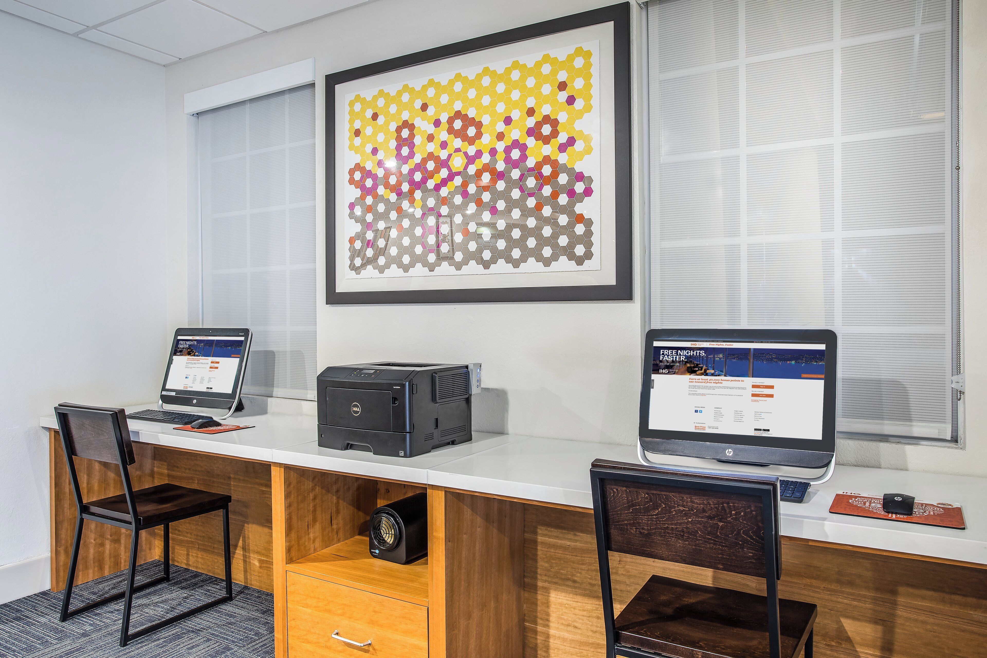 A modern office space with two desks, each with a laptop and a desktop computer. There is a framed abstract art piece on the wall and a window with blinds.