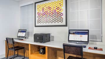 A modern office space with two desks, each with a laptop and a desktop computer. There is a framed abstract art piece on the wall and a window with blinds.