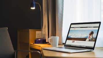 Um laptop sobre uma mesa de madeira com uma caneca branca, uma luminária e uma tomada em um quarto de hotel.
