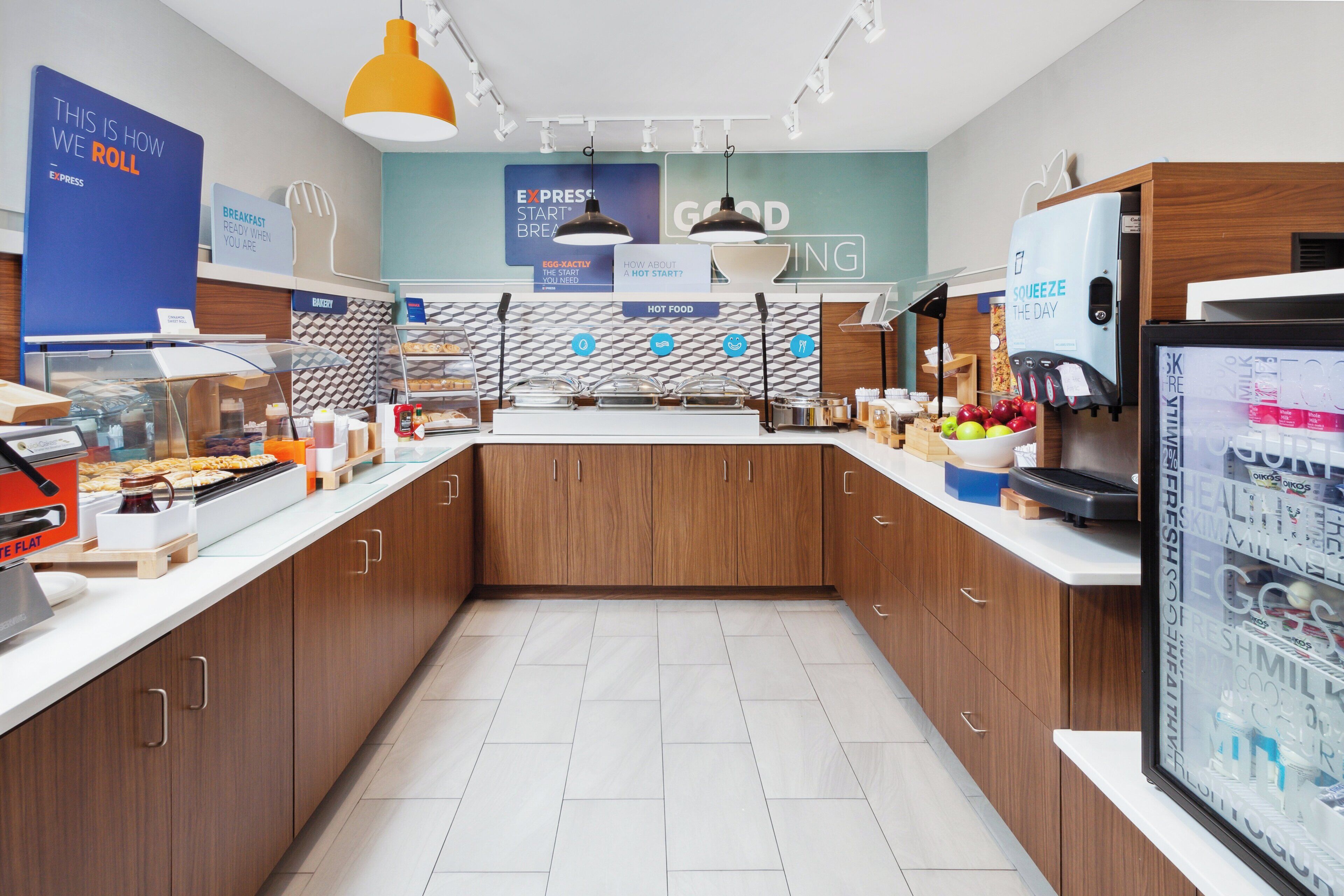 A modern breakfast area with a counter displaying various food items, a coffee machine, and a refrigerator with fresh produce.