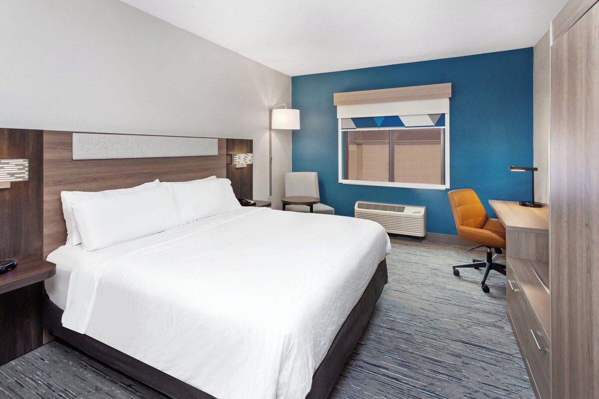 Une chambre d’hôtel avec un grand lit, un bureau, un fauteuil de bureau, une lampe et une fenêtre avec des stores.