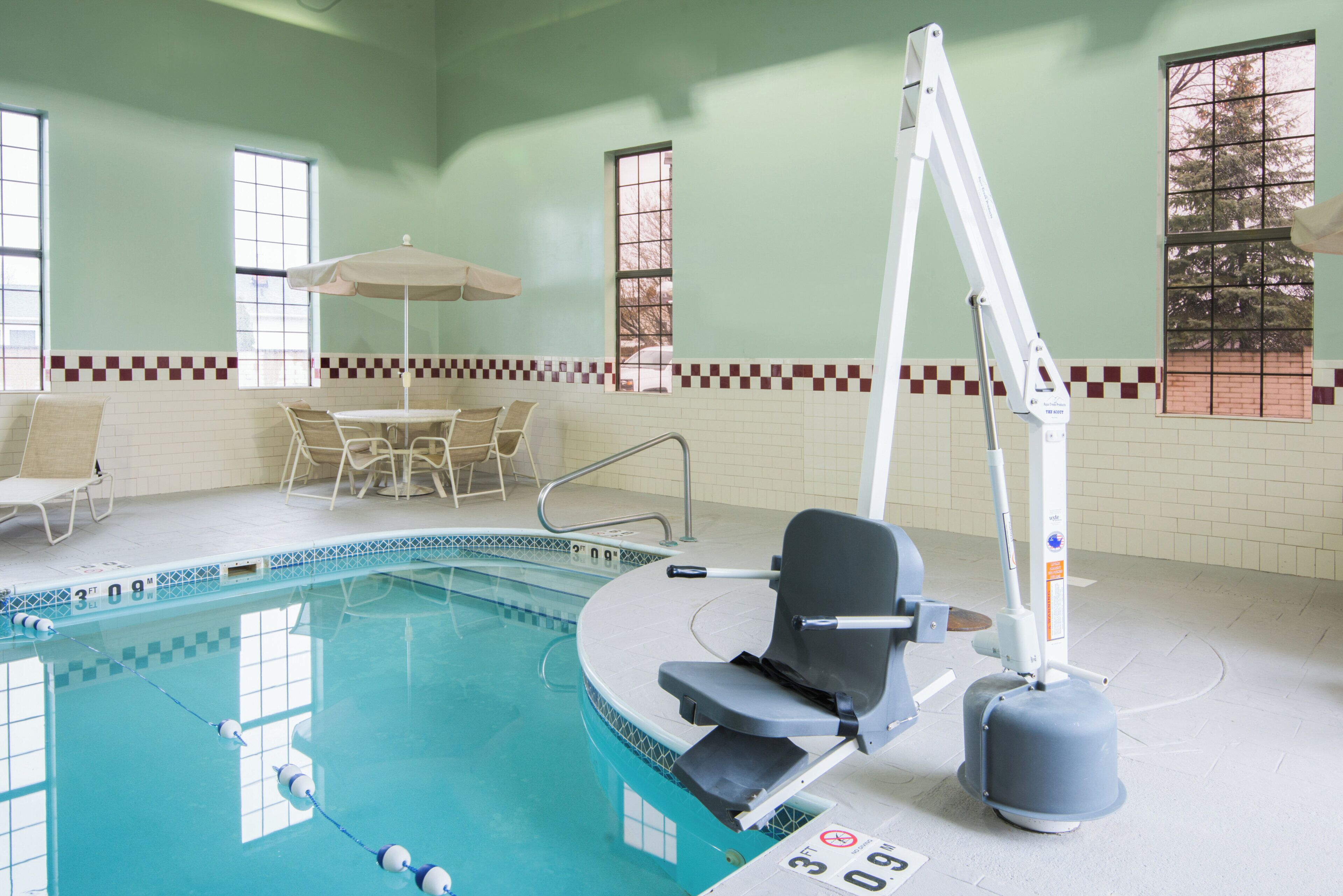 An indoor swimming pool with a pool lift chair, lounge chairs, and a table with an umbrella.